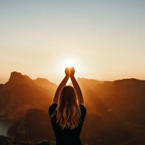 women sitting on a cliff, looking into the sunset with her hands clasped together raised above her depicting past life regression has occurred